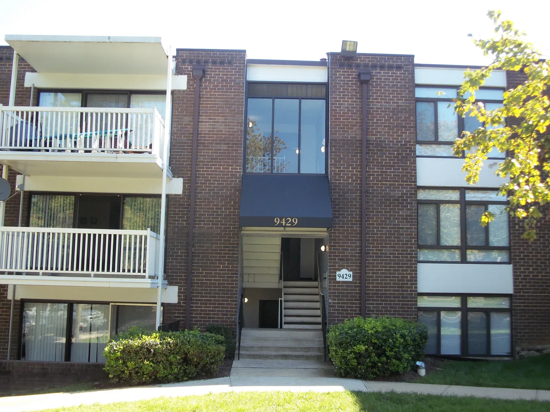 Apartment building entrance with brick facade, balconies, and central stairs leading to glass doors