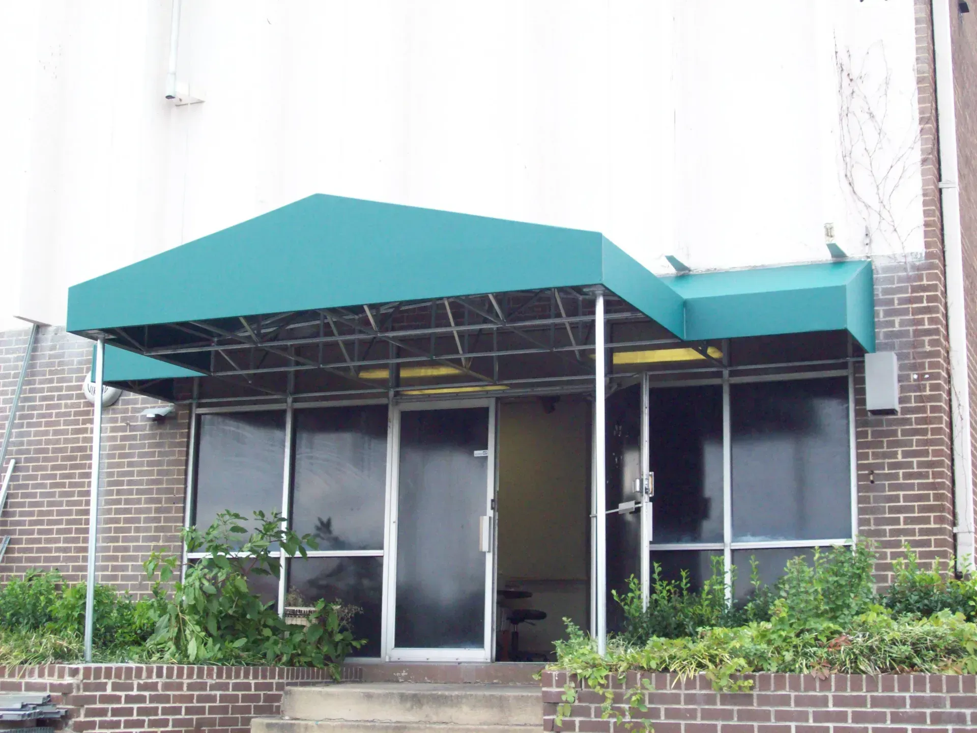 Building entrance with teal awning, glass doors, and small shrubs in front