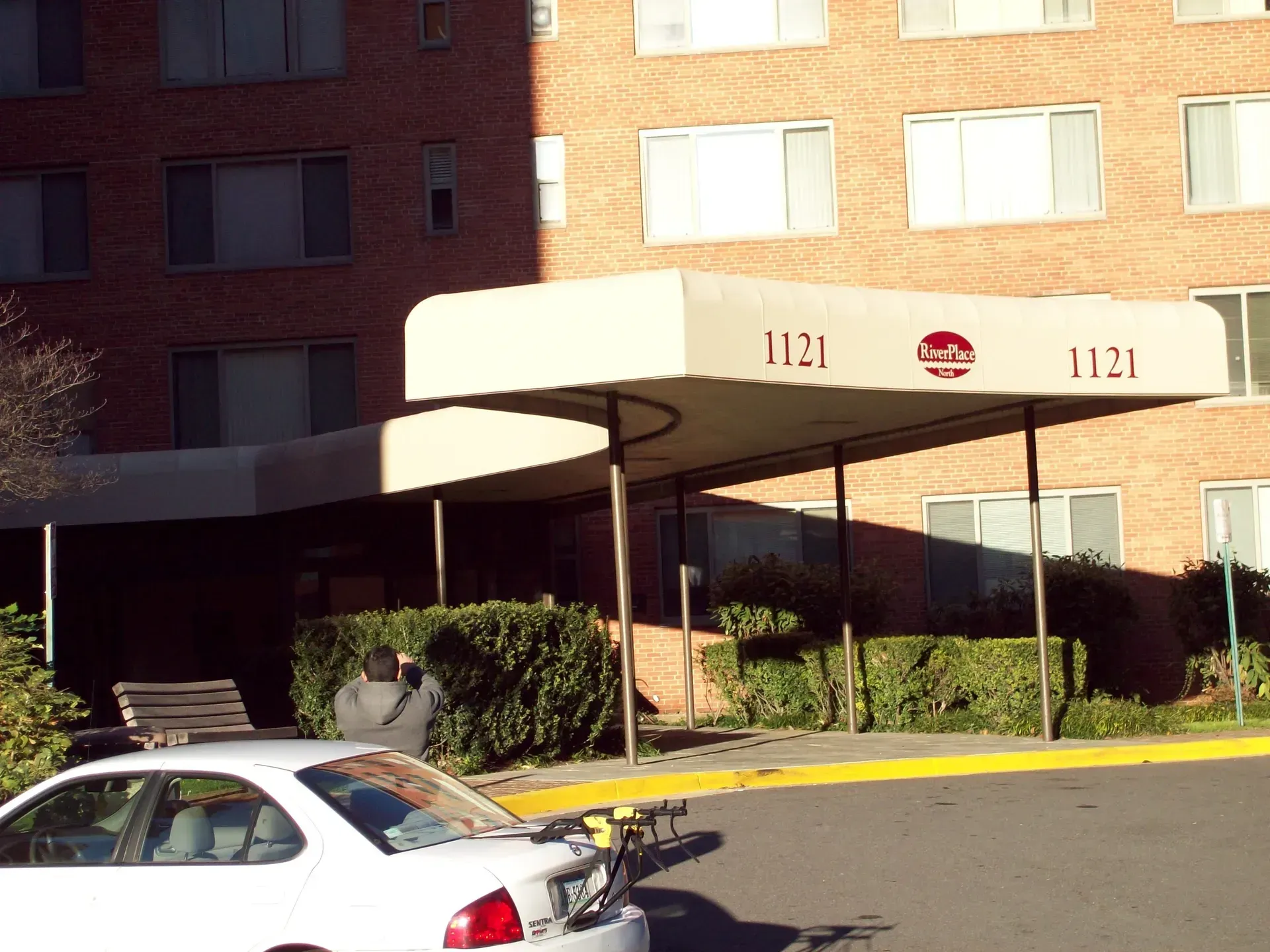 Apartment building entrance with awning, benches, and a parked white car in front