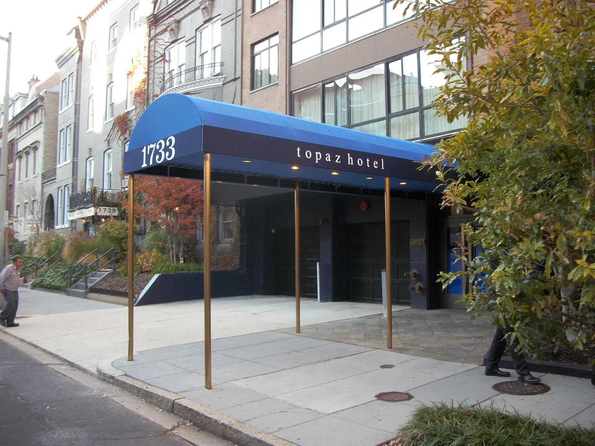 Blue-awning entrance to a modern building on a city sidewalk, with trees and a few pedestrians nearby