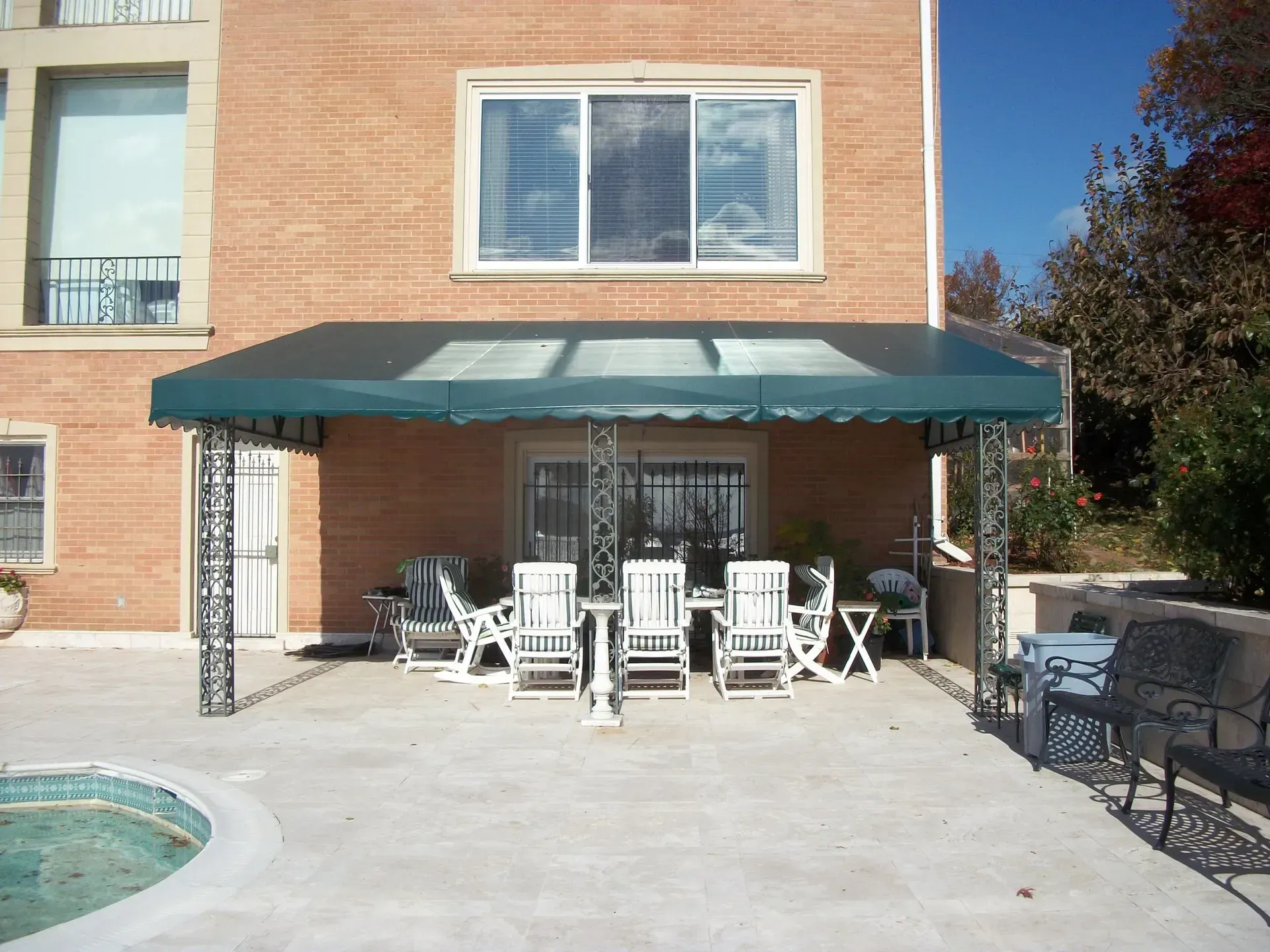 Backyard patio with green awning, white patio chairs, and a pool beside a brick house.