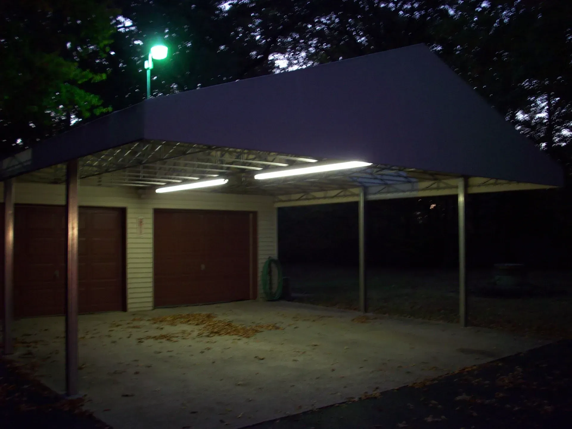 Dimly lit outdoor carport with metal roof, concrete floor, and closed red garage doors at night