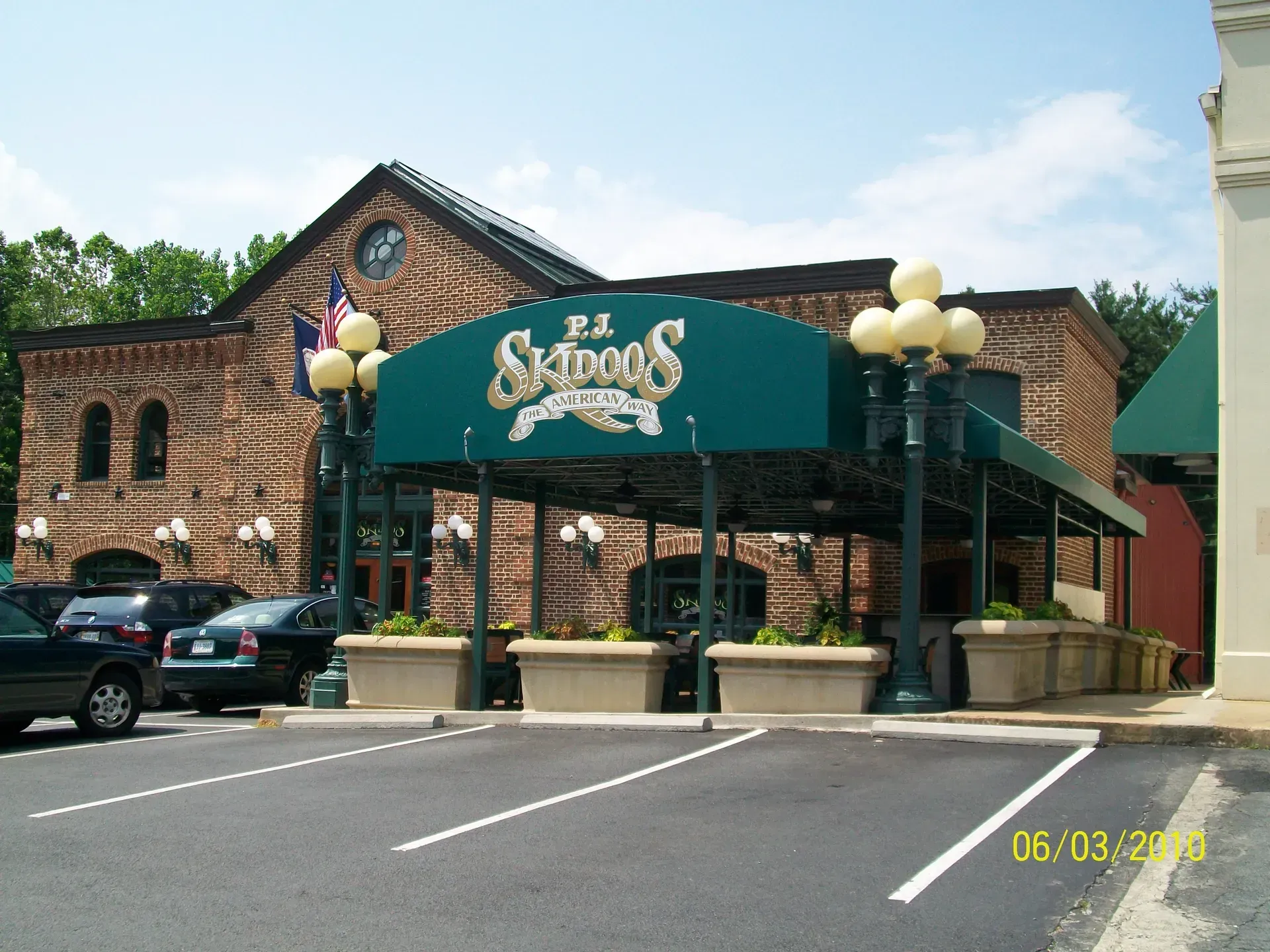 Brick restaurant entrance with green awning, parking lot, and parked cars on a sunny day