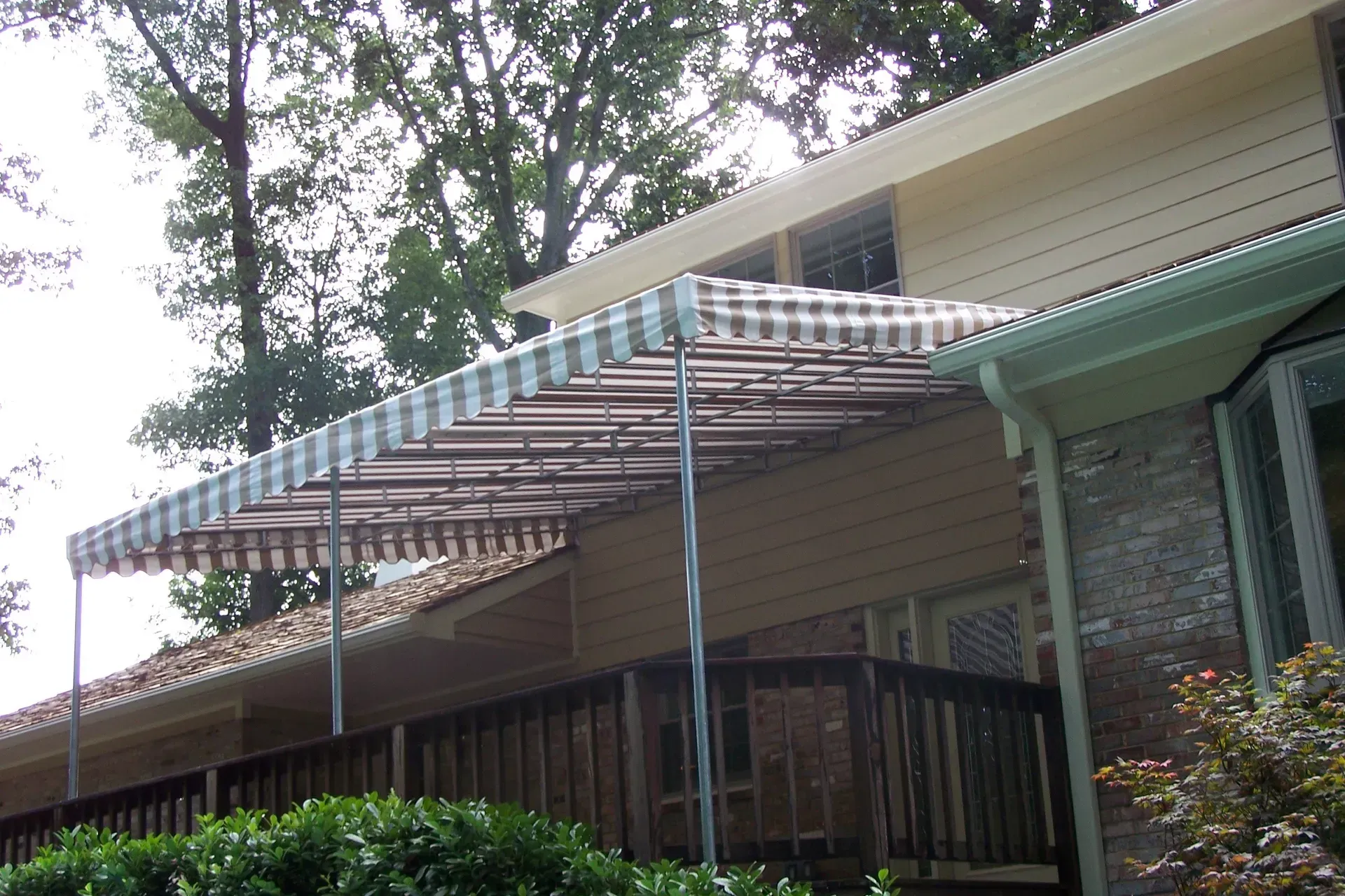 Striped patio awning attached to a house, shading a porch with wooden railings and trees overhead