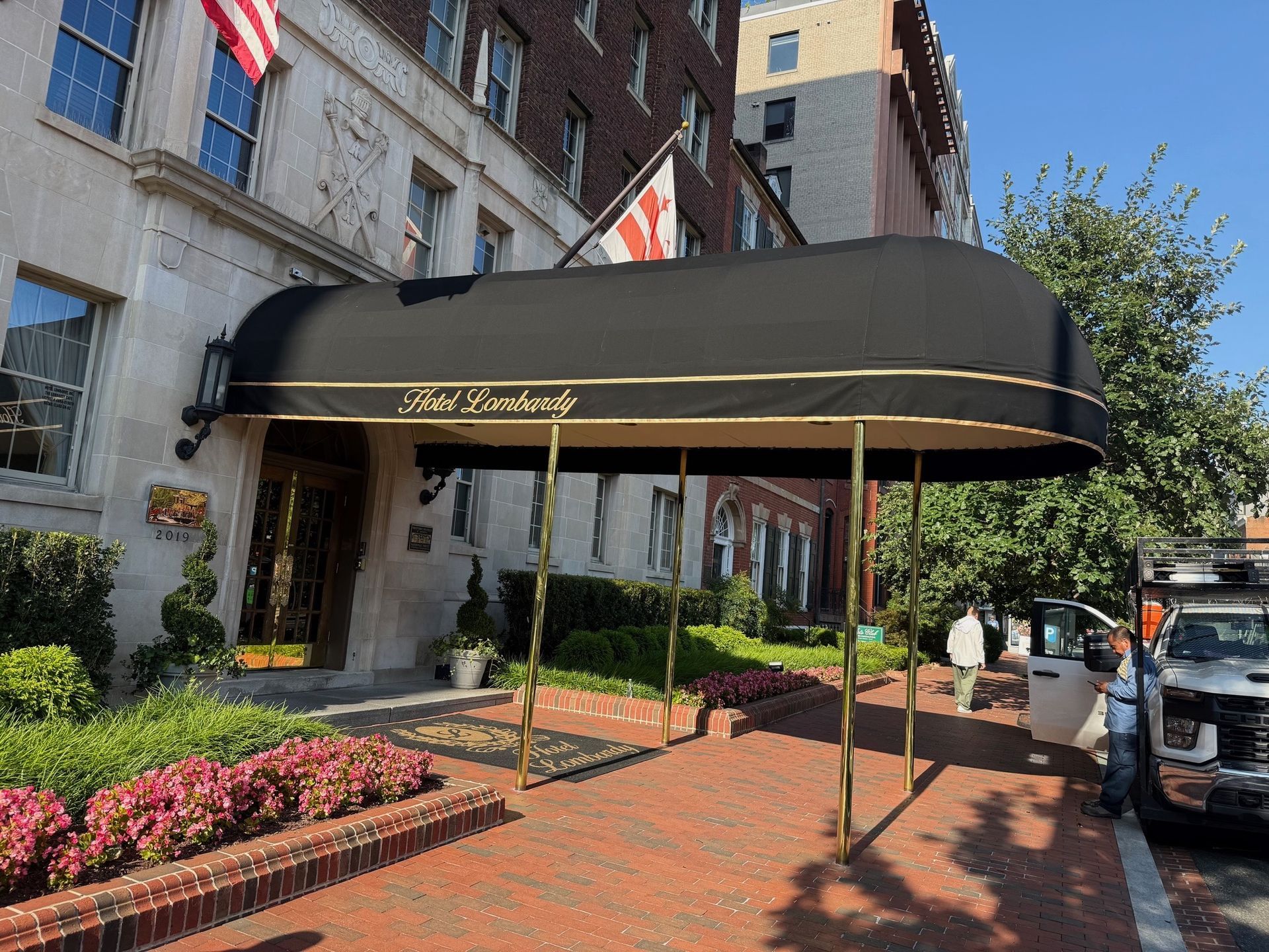 Hotel entrance with black awning, red brick walkway, flower beds, and parked cars outside a building.