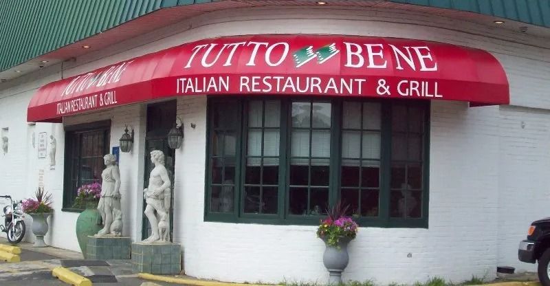 Red awning reading “Tuto Bene Italian Restaurant & Grill” on a white storefront with windows and statues