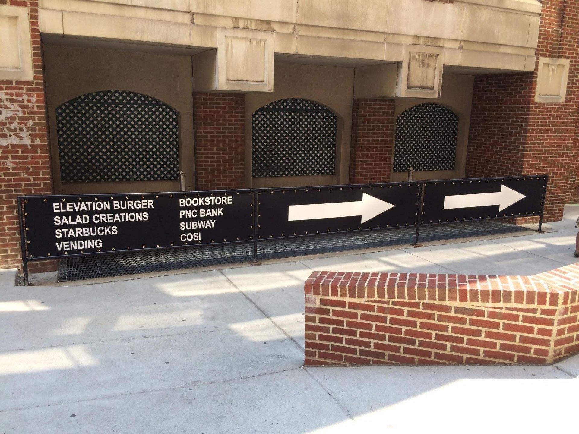 Black directional signs with white arrows on a brick building walkway, with a low brick planter in front.