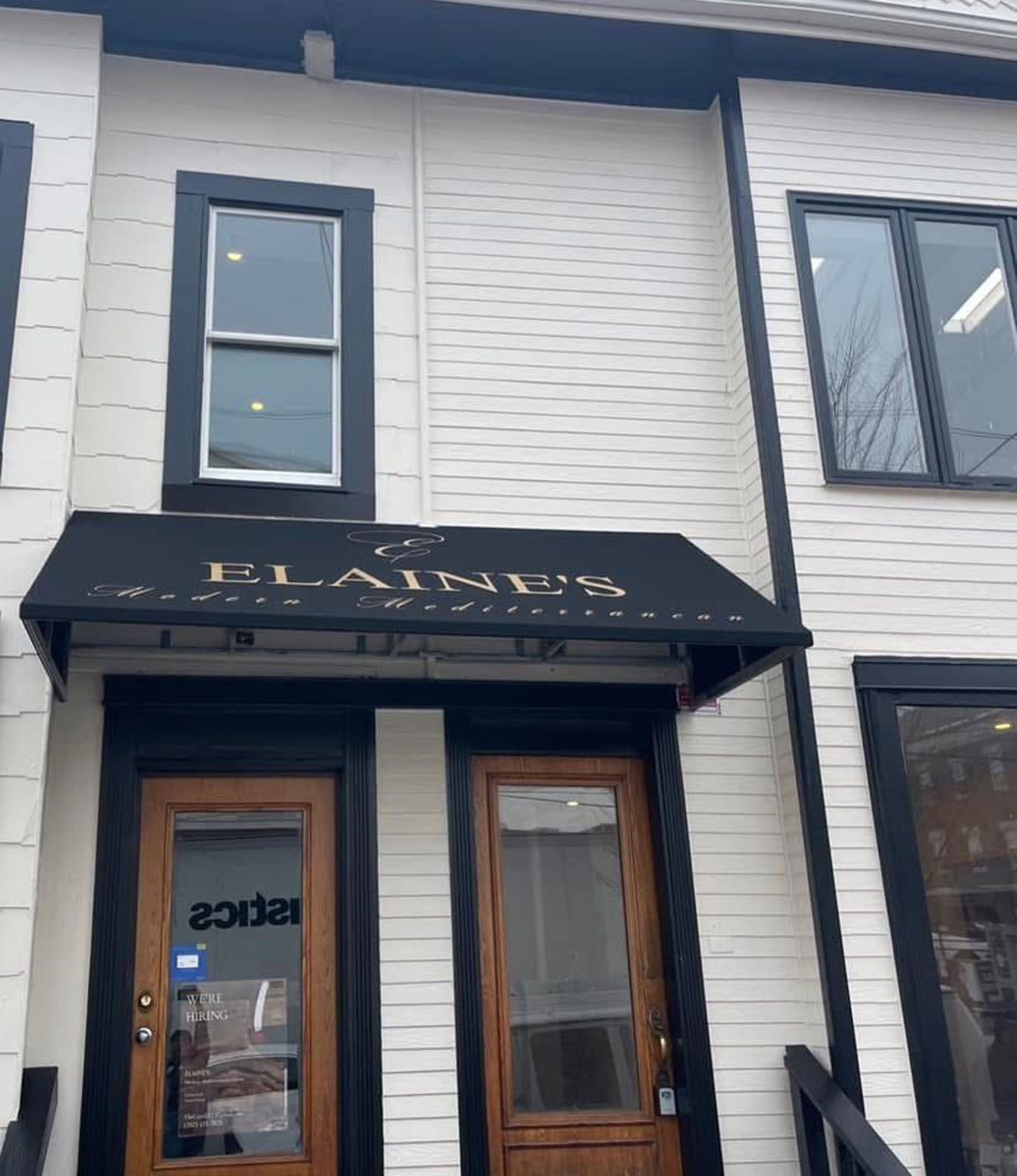 Storefront with black awning, white siding, and wooden double doors on a street.