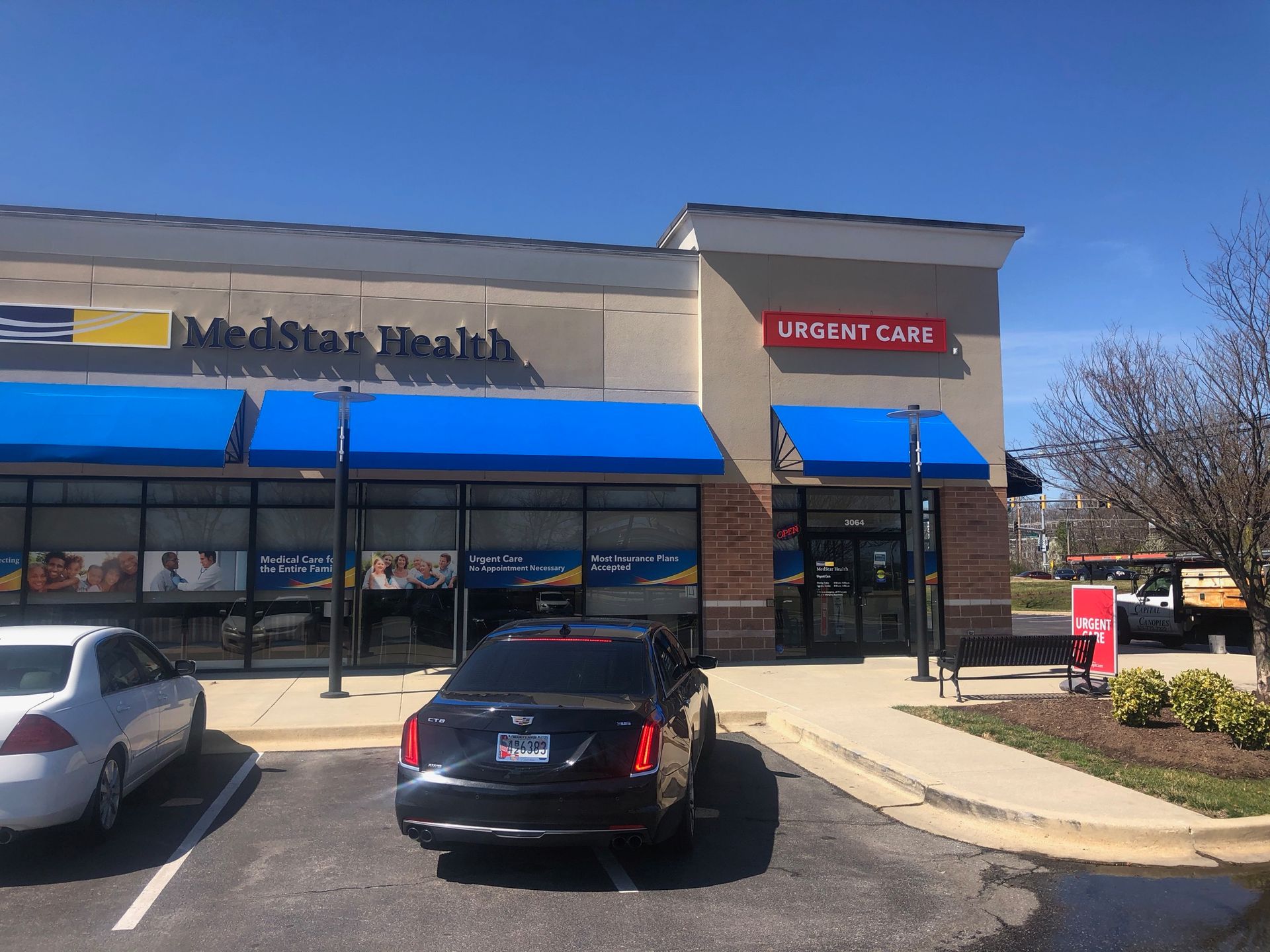 Shopping center storefronts with blue awnings, parked cars, and a clear blue sky.