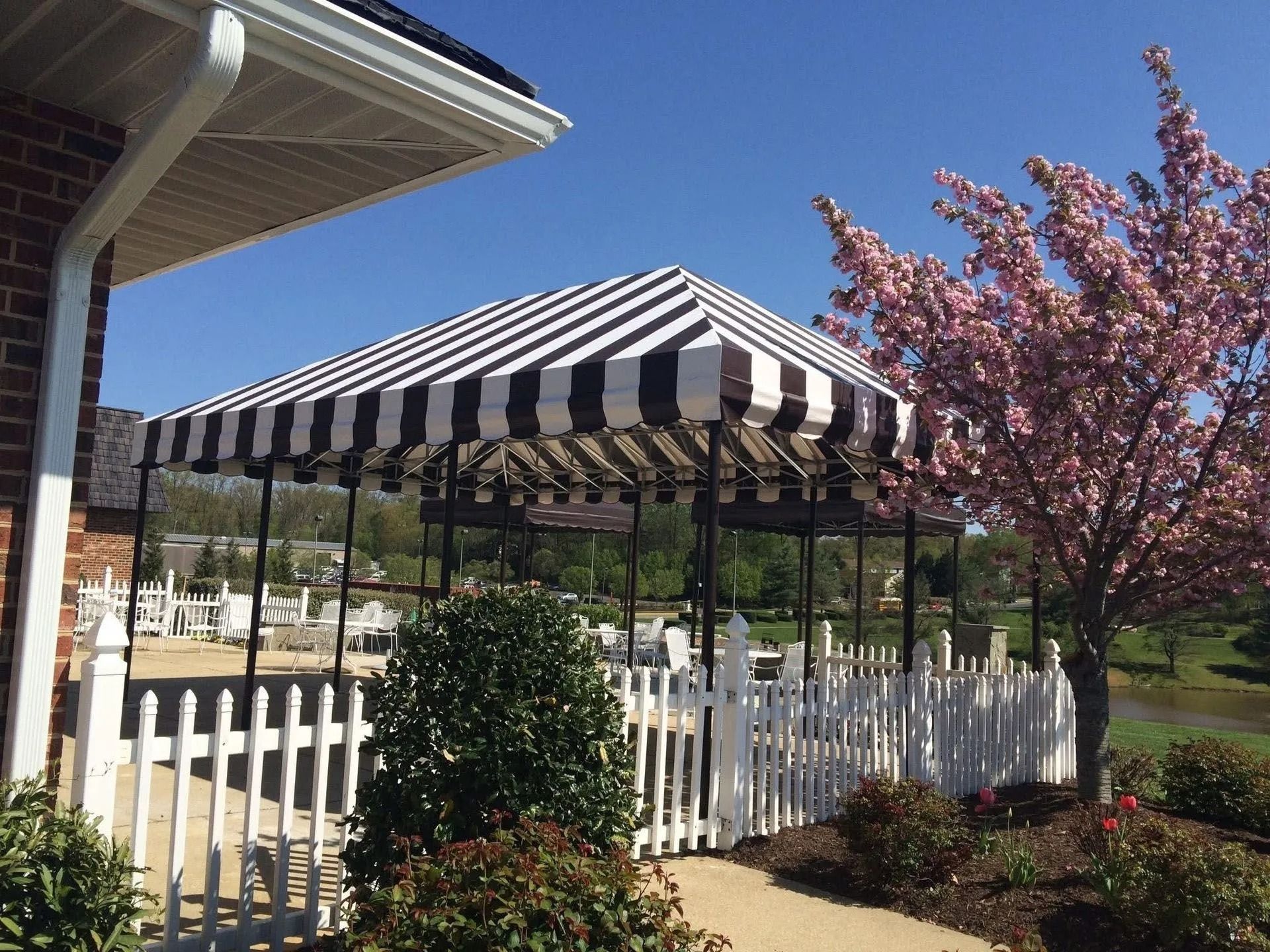 Striped patio awning beside a white picket fence and pink flowering tree in a sunny yard