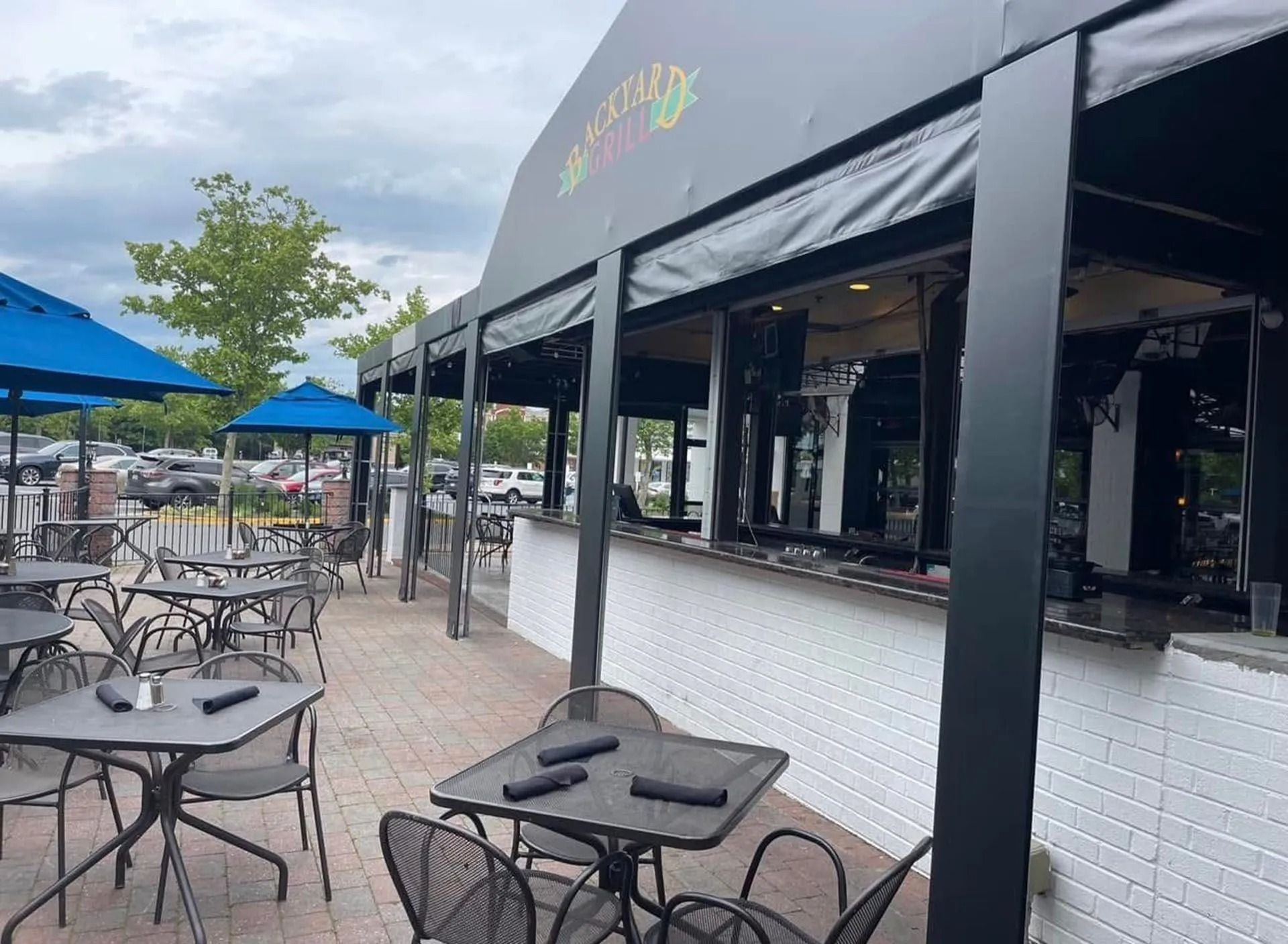 Outdoor café seating with black tables and chairs beside a white building and blue umbrellas