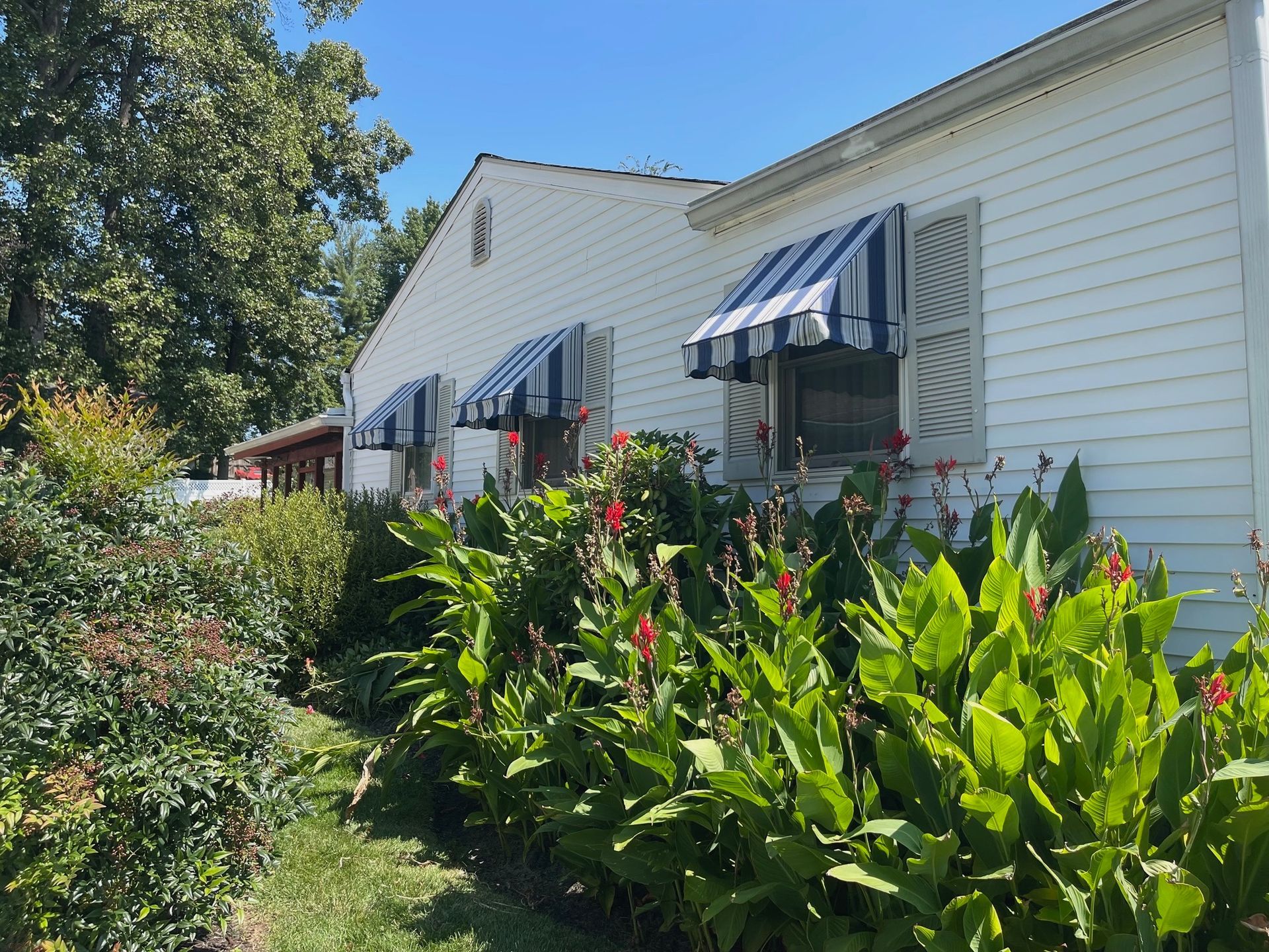 White house with blue-striped awnings and lush green shrubs in the foreground on a sunny day.