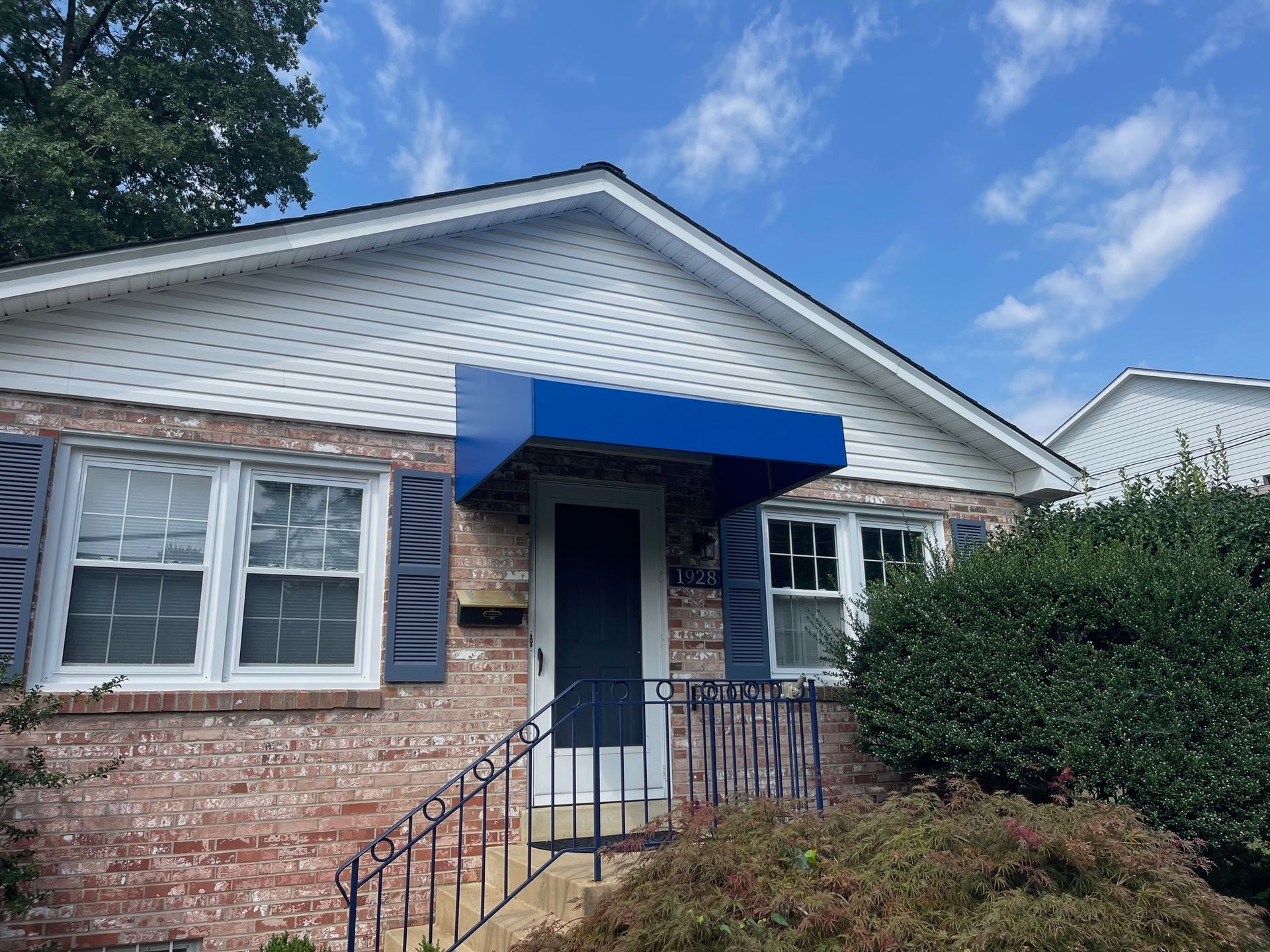 Brick house entry with blue awning, white-trimmed windows, and a front porch under a cloudy sky.