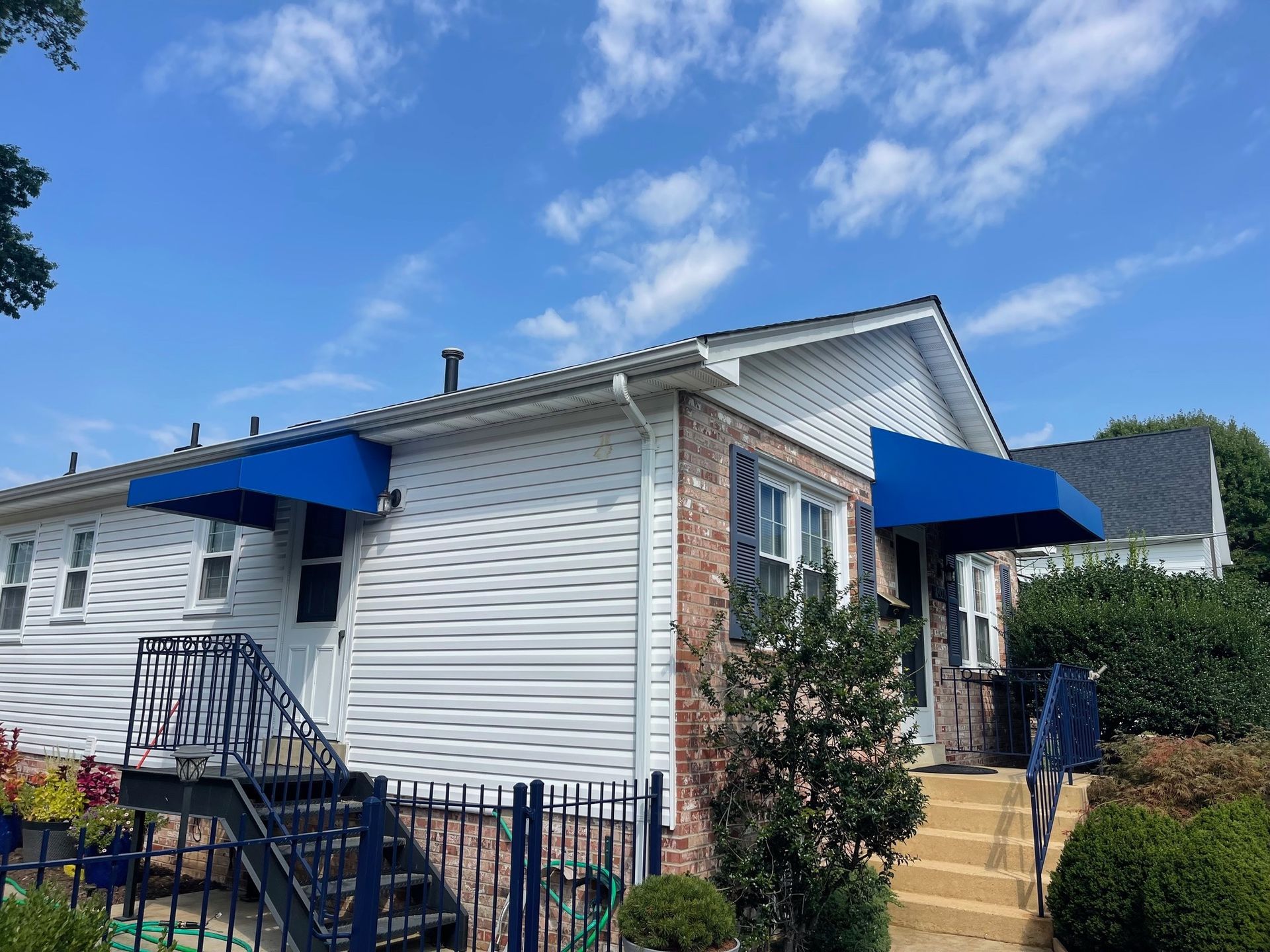 White house with blue awnings, front steps, and a small garden under a bright blue sky.