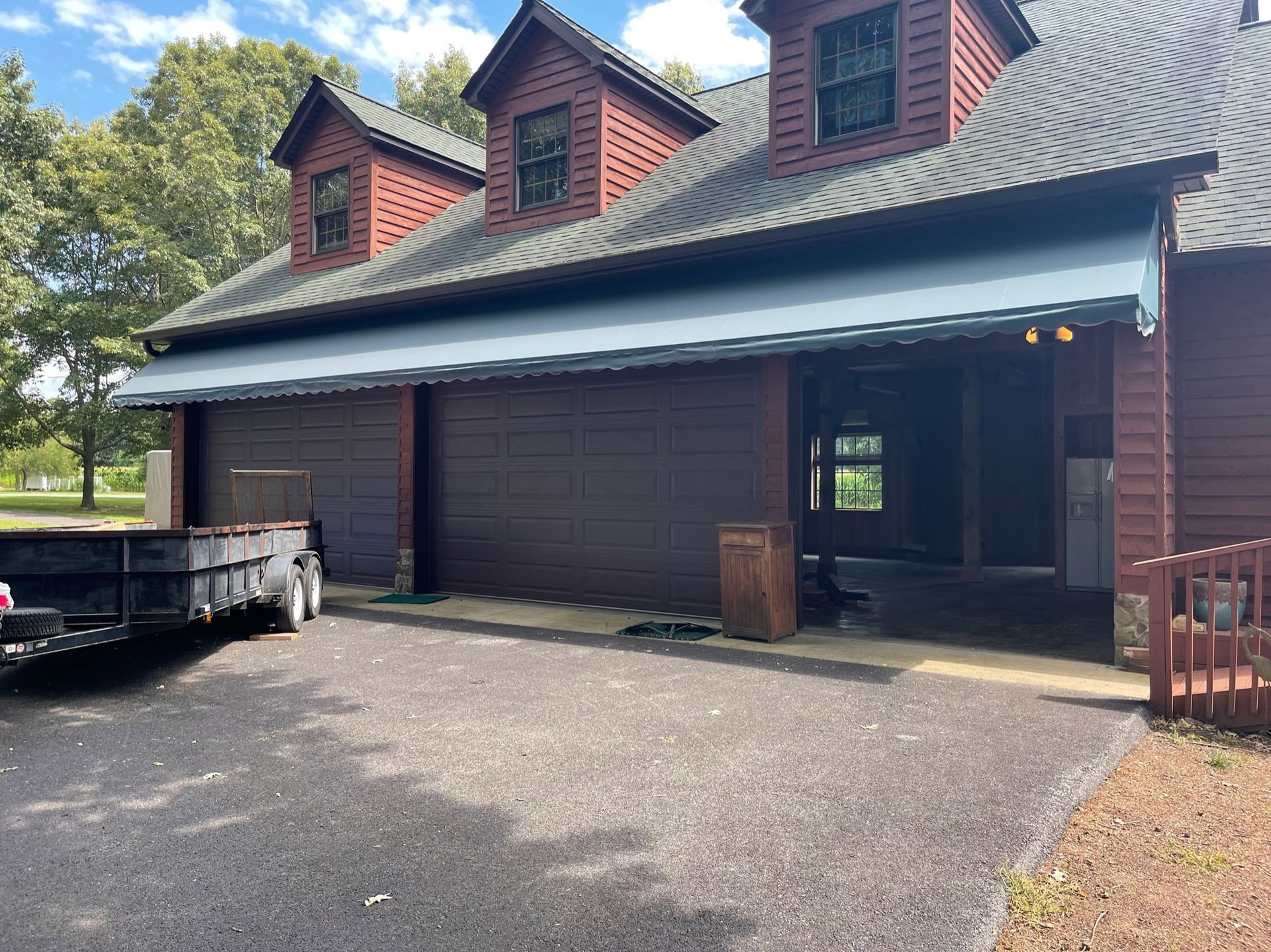 Brown cabin-style building with red dormers, a carport, and a trailer in a paved lot under trees.