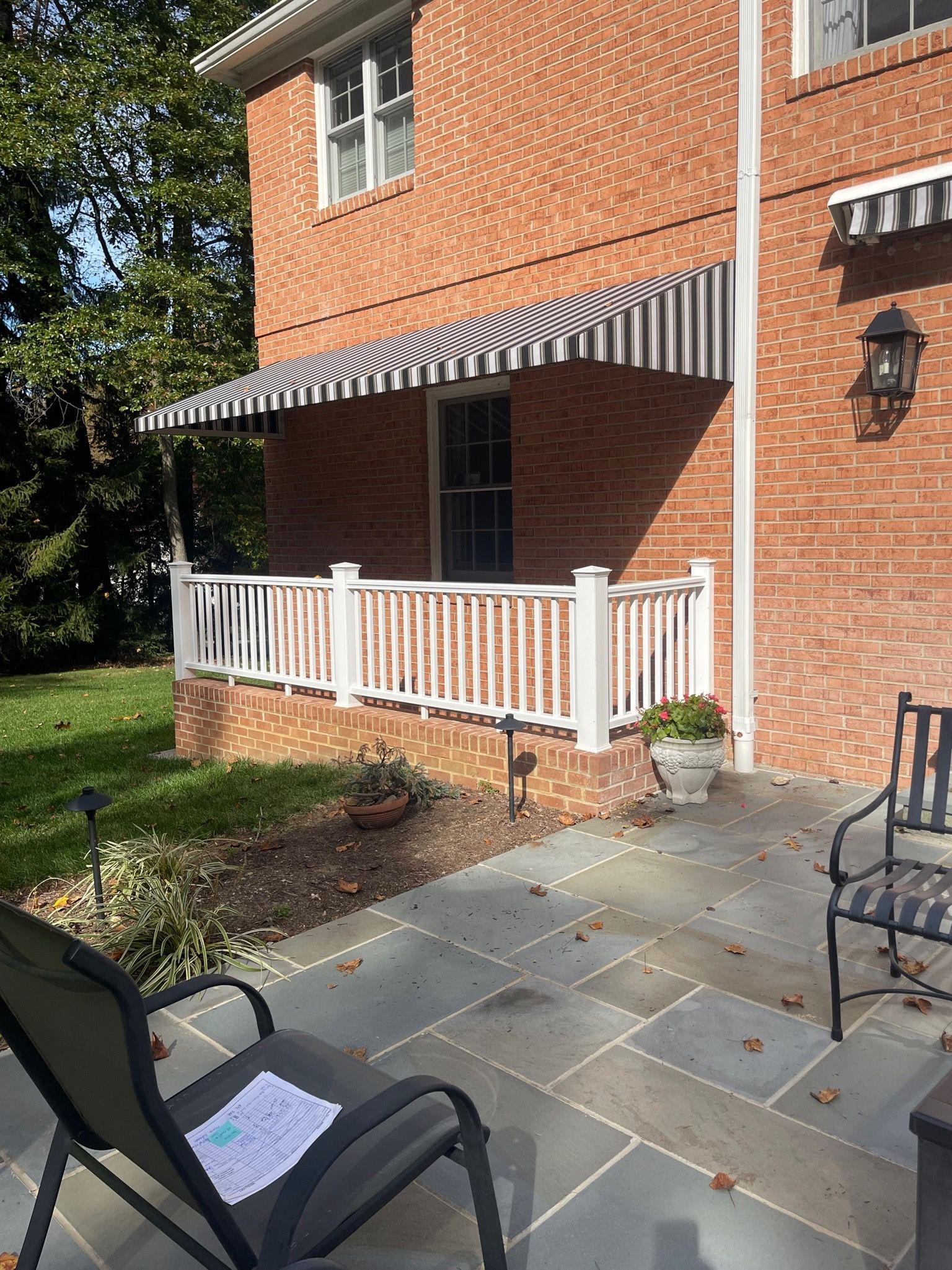 Brick house patio with white railing, striped awning, and outdoor chairs in a sunny yard.