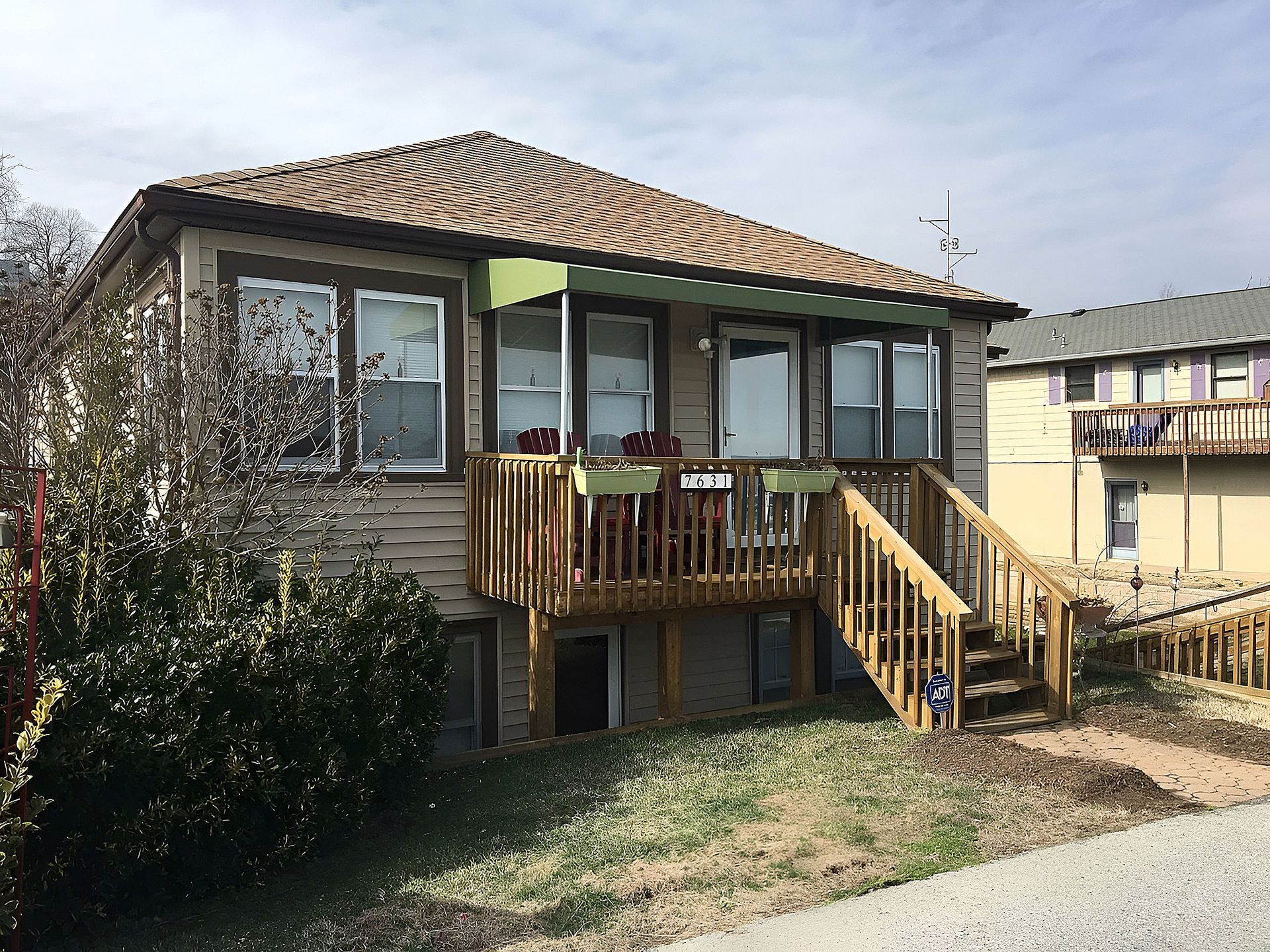 Small elevated house with a tiled roof, front balcony, stairs, and a yard on a sunny day.