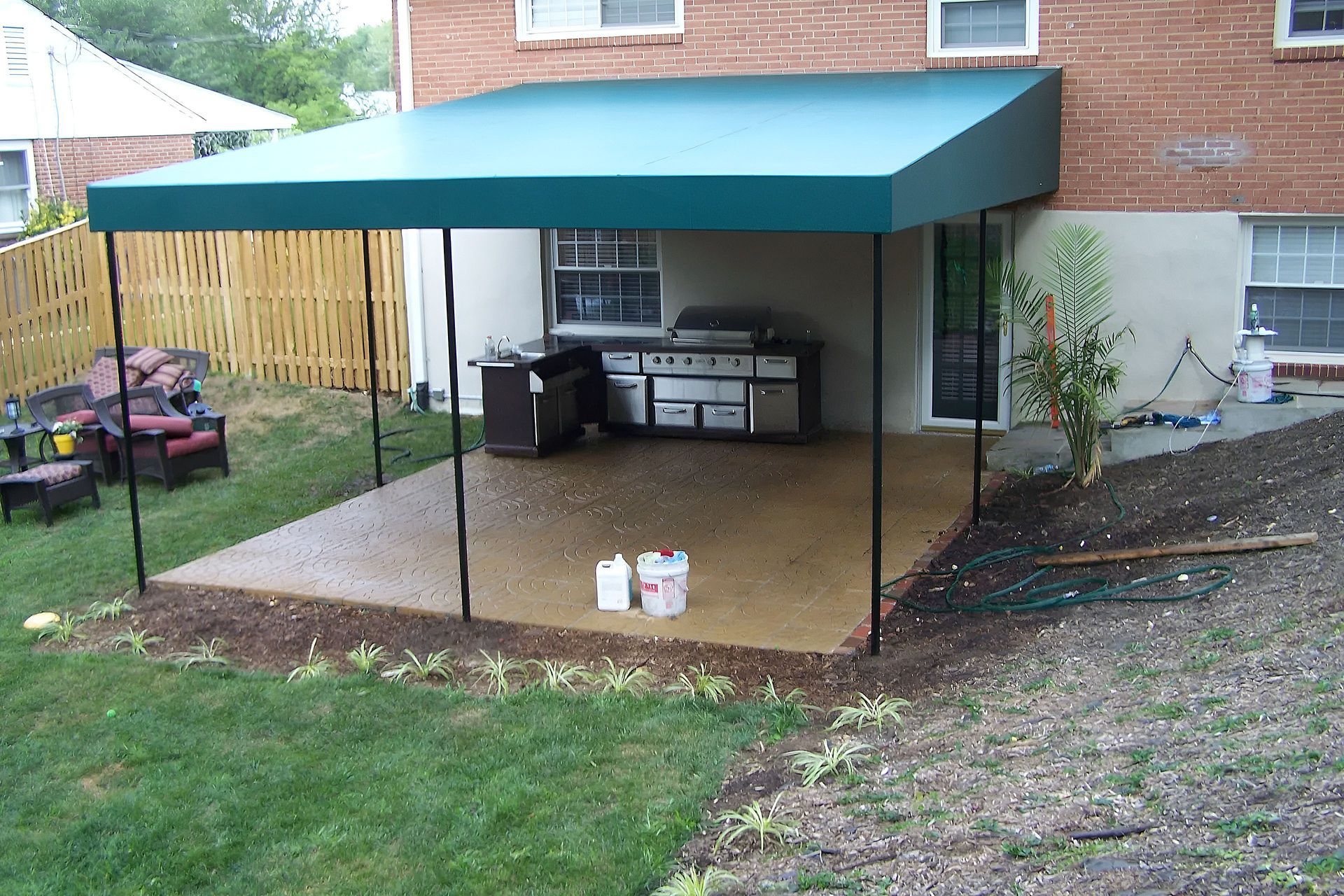 Backyard patio with a teal awning, grill, and small storage setup beside a brick house.