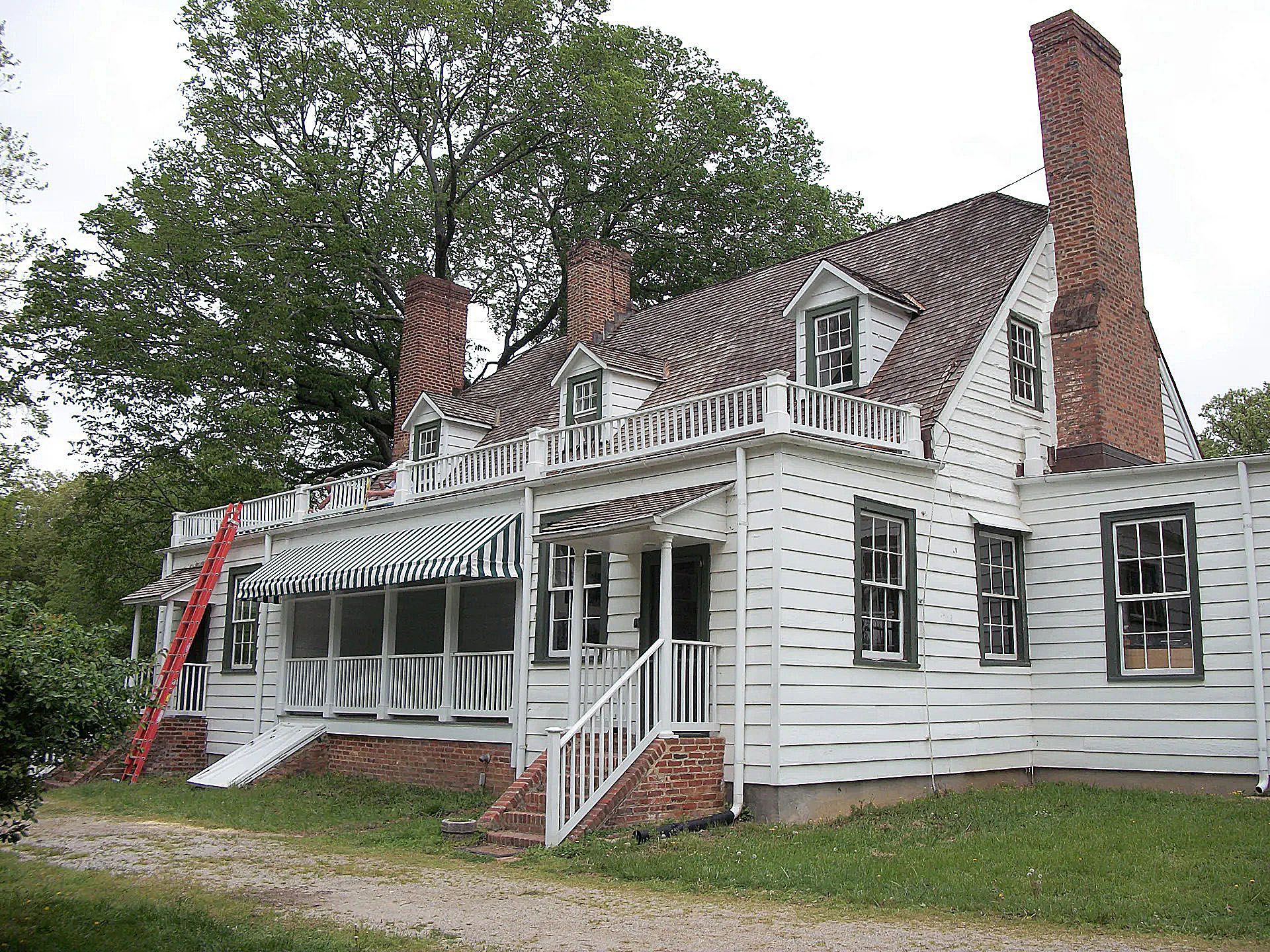 White house with porch, red ladder, and brick chimney on a grassy lot amid trees.