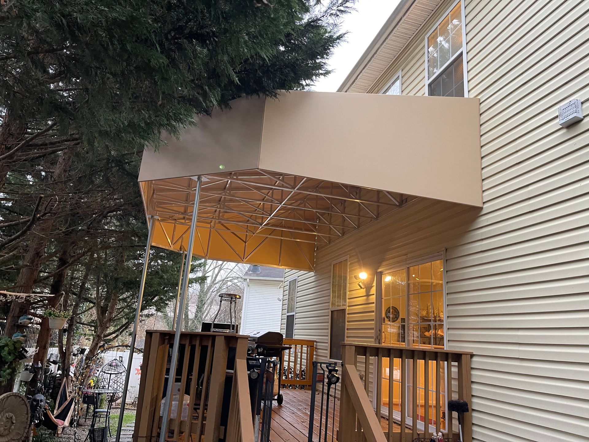 Covered outdoor entrance with beige awning, wooden deck, and lights beside a house.