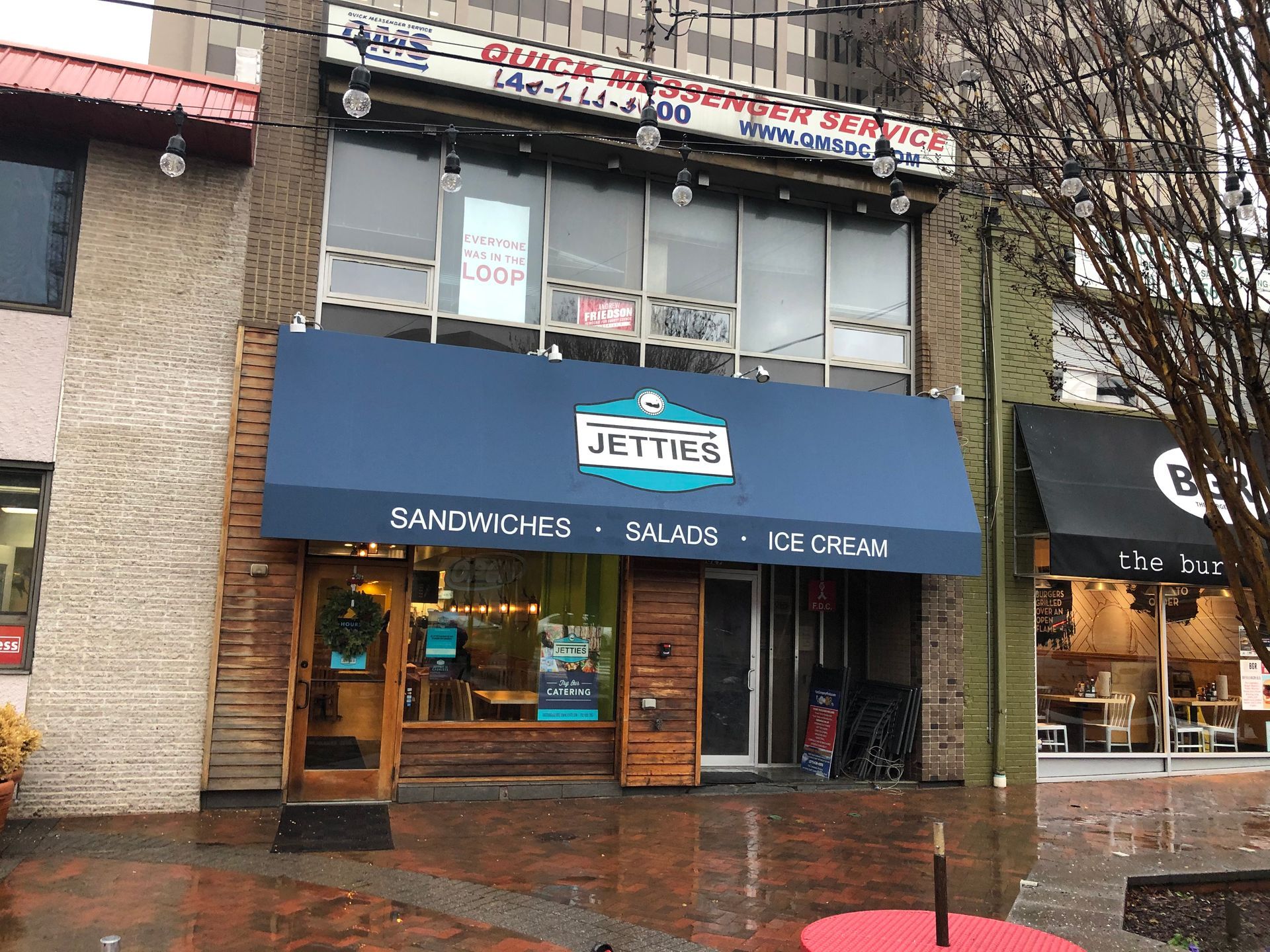 Storefront with a blue awning on a rainy street, reflected in the wet pavement.
