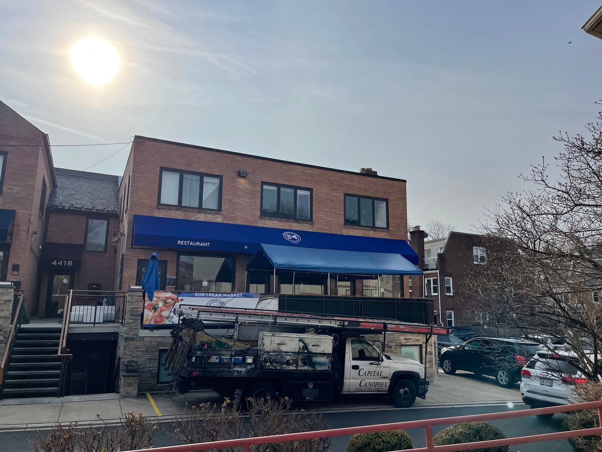 Street view of a brick storefront with a blue awning, parked vehicles, and steps under a bright sun.