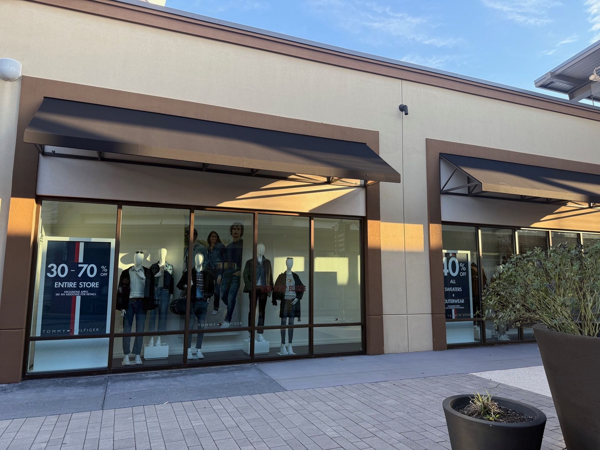 Outdoor shopping center storefront with large glass windows, awnings, and sale signs under a bright sky.
