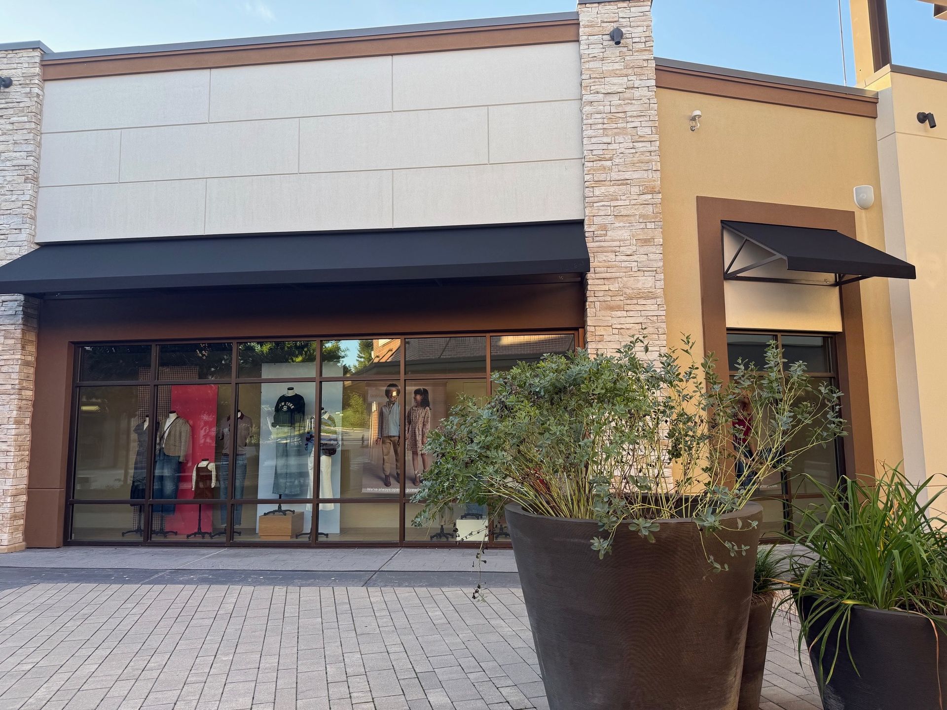 Storefront with glass windows, black awnings, stone columns, and large planters on a paved walkway.