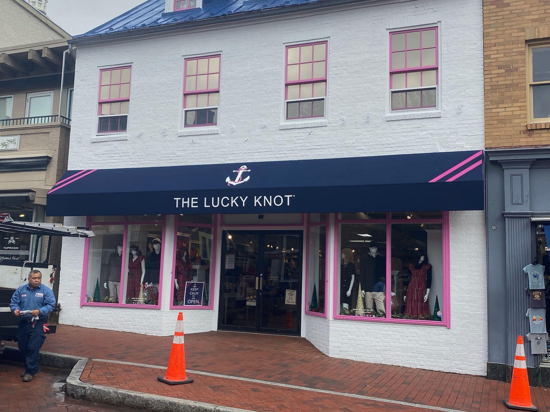 Storefront of “The Jockey Knot” with navy awning and pink window trim on a wet street, orange cones out front.