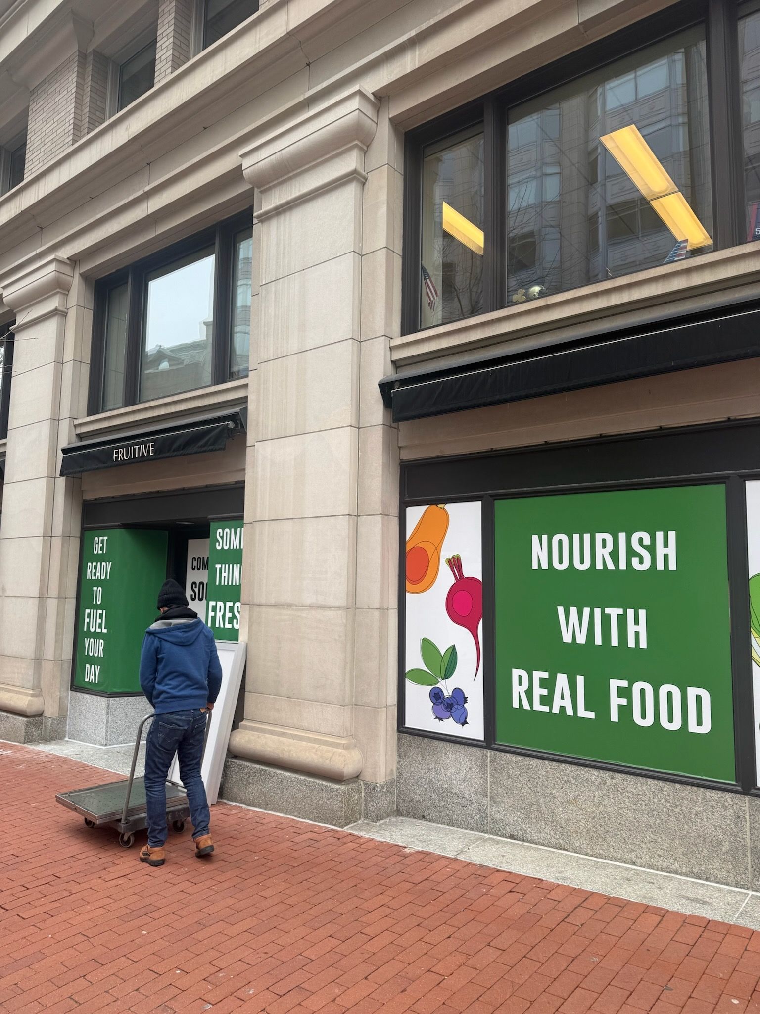 Person standing outside storefront with green sign reading “NOURISH WITH REAL FOOD”.