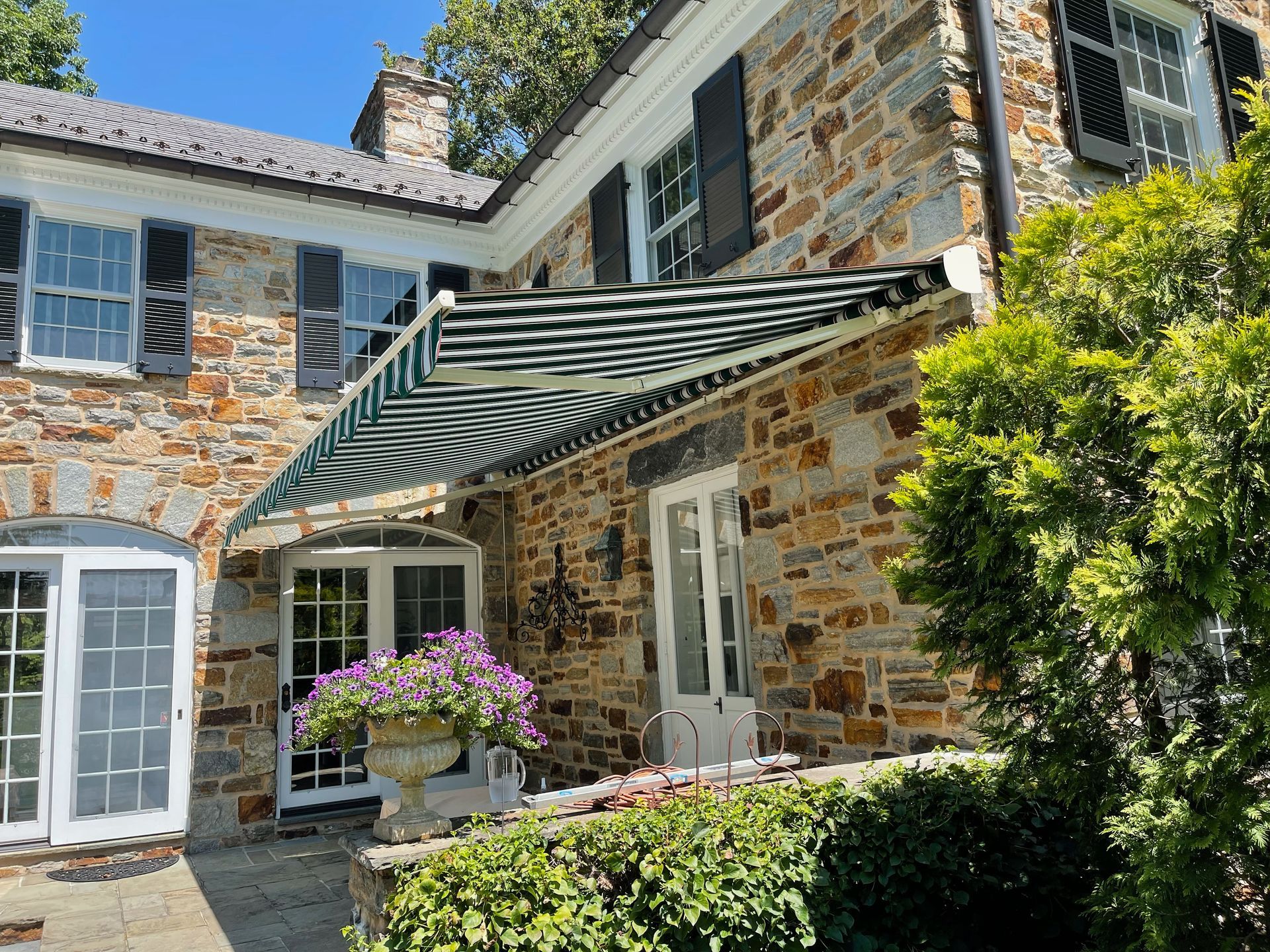 Stone house courtyard with striped awning, white doors, flowering plants, and blue shutters.