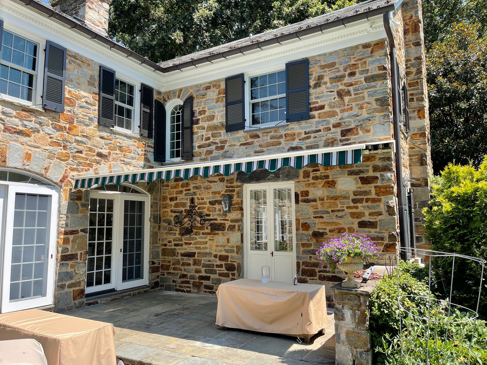 Stone house patio with white-framed windows, striped awning, and covered outdoor furniture.