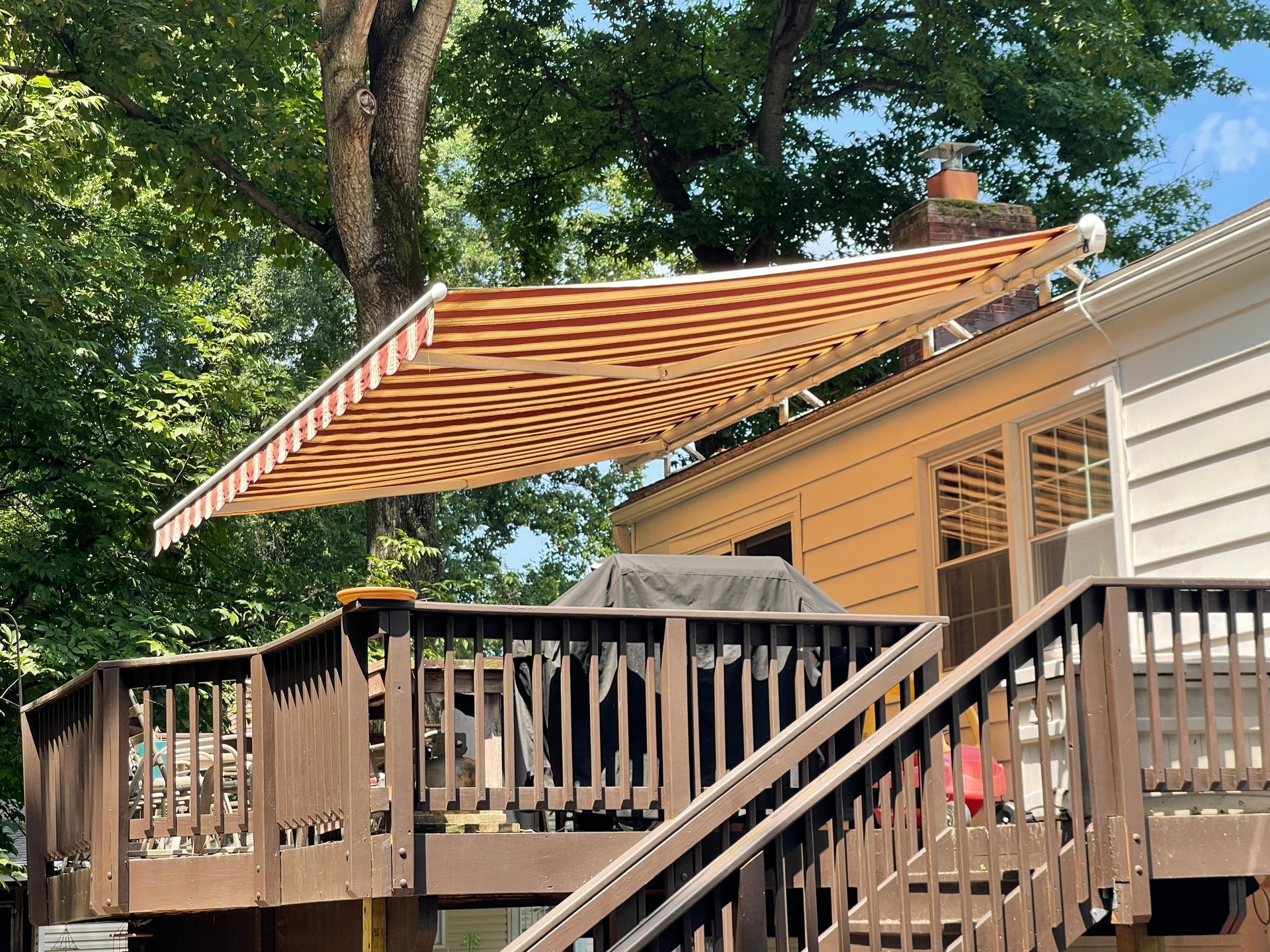 Back deck with striped retractable awning beside a house, framed by trees and wooden railing.
