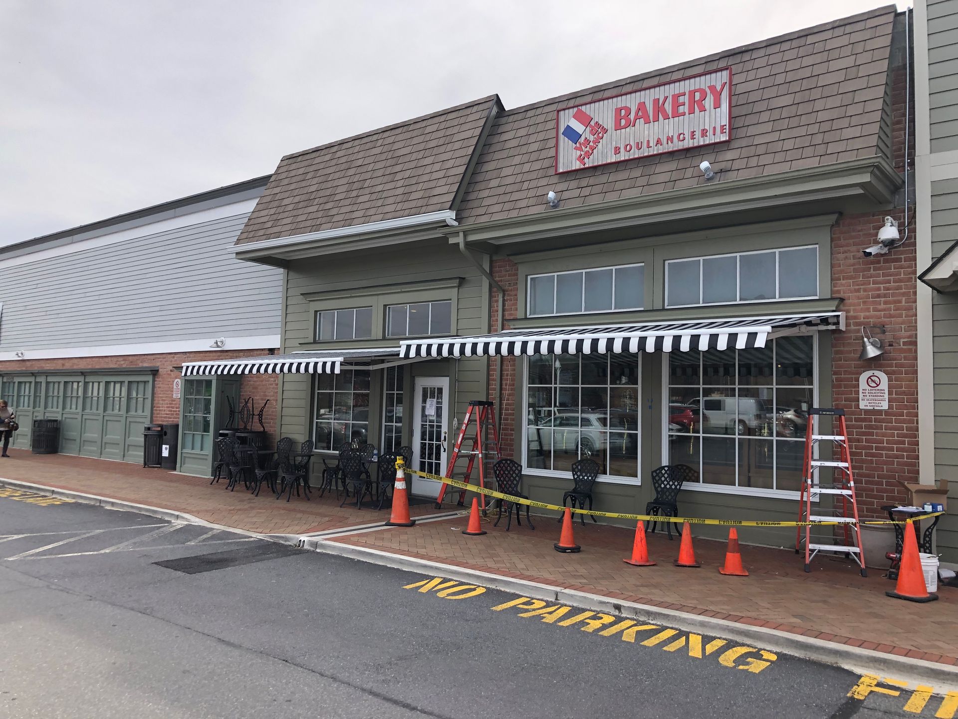 Bakery storefront with striped awning, orange cones, and a ladder outside in a parking lot.