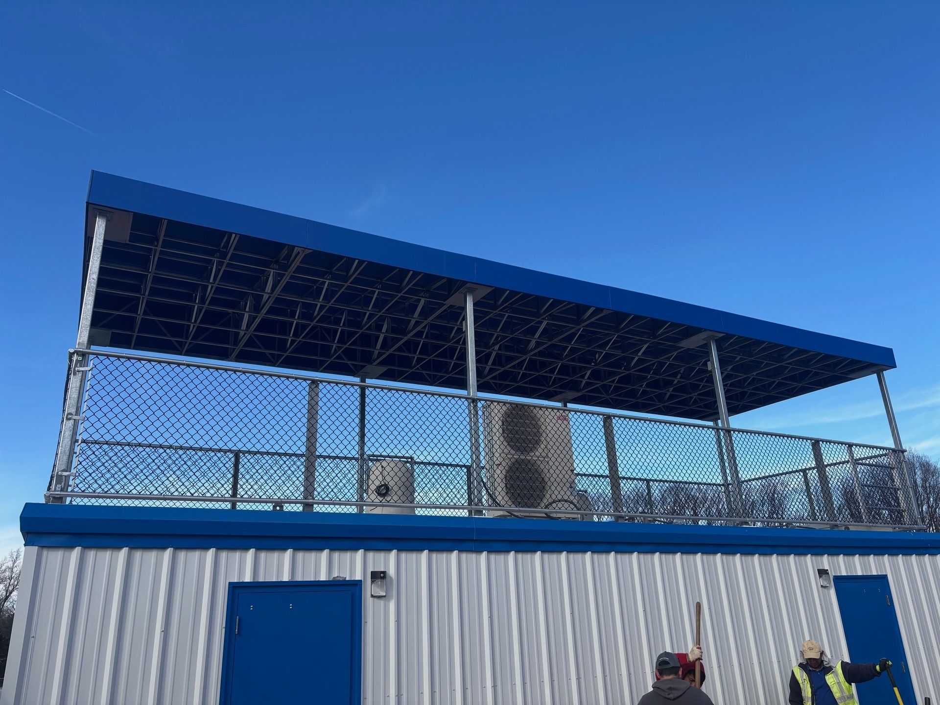 Blue metal building with a fenced rooftop deck under a clear sky, with a blue door below.