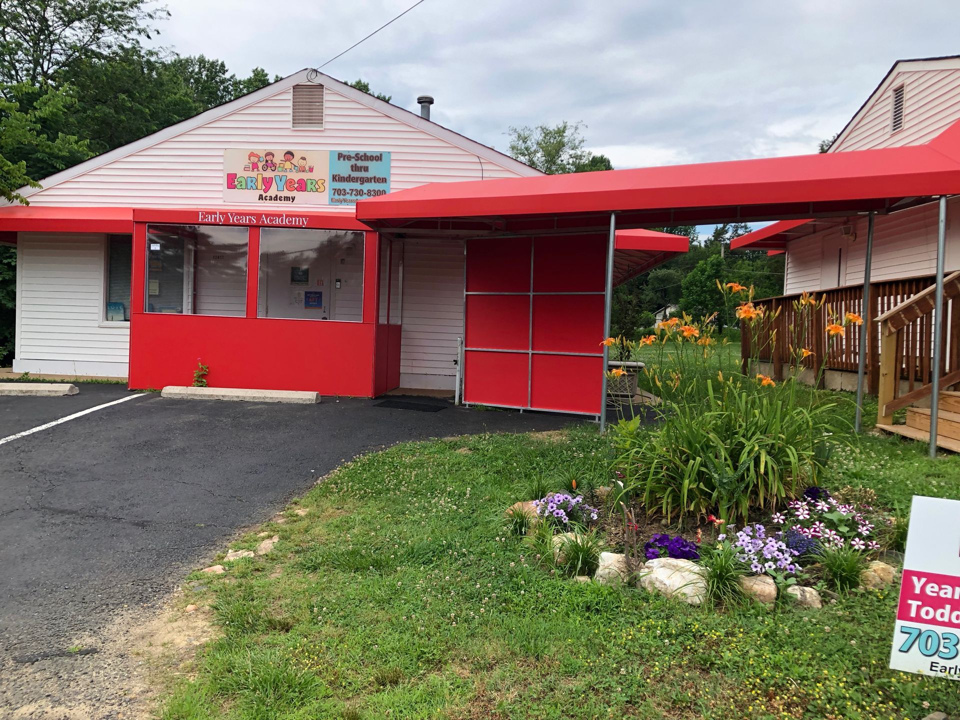 Red roadside produce stand with white roof, red awning, and flower bed beside a gravel driveway.