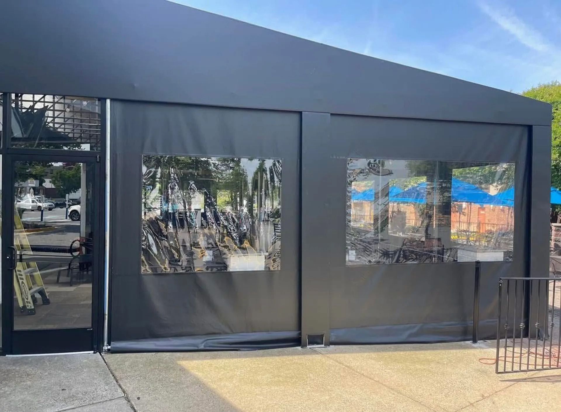Black outdoor structure with tinted windows and a dark canopy on a sunny sidewalk.