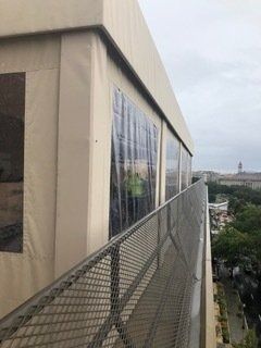 Balcony walkway beside beige building with mesh railing, overlooking a distant cityscape under cloudy sky