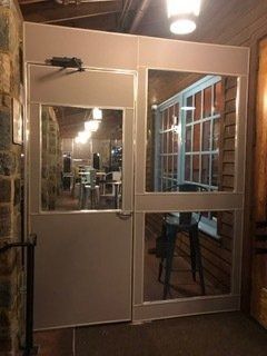 Glass and metal entry door at night, with lights and a chair visible inside.