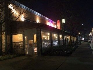 Nighttime street view of a lit restaurant with large windows and a sidewalk outside