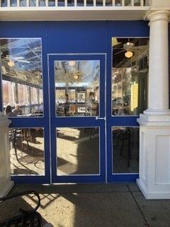 Blue restaurant entrance with glass double doors under white columns, showing chairs and tables inside.