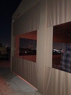 Nighttime exterior of a tent-like structure with large window openings and city lights in the background