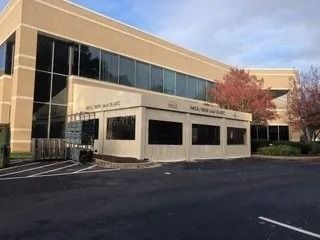 Modern office building with beige and glass exterior, black parking lot, and a red tree nearby