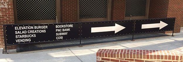 Black directional sign with white text and arrows outside a brick building.