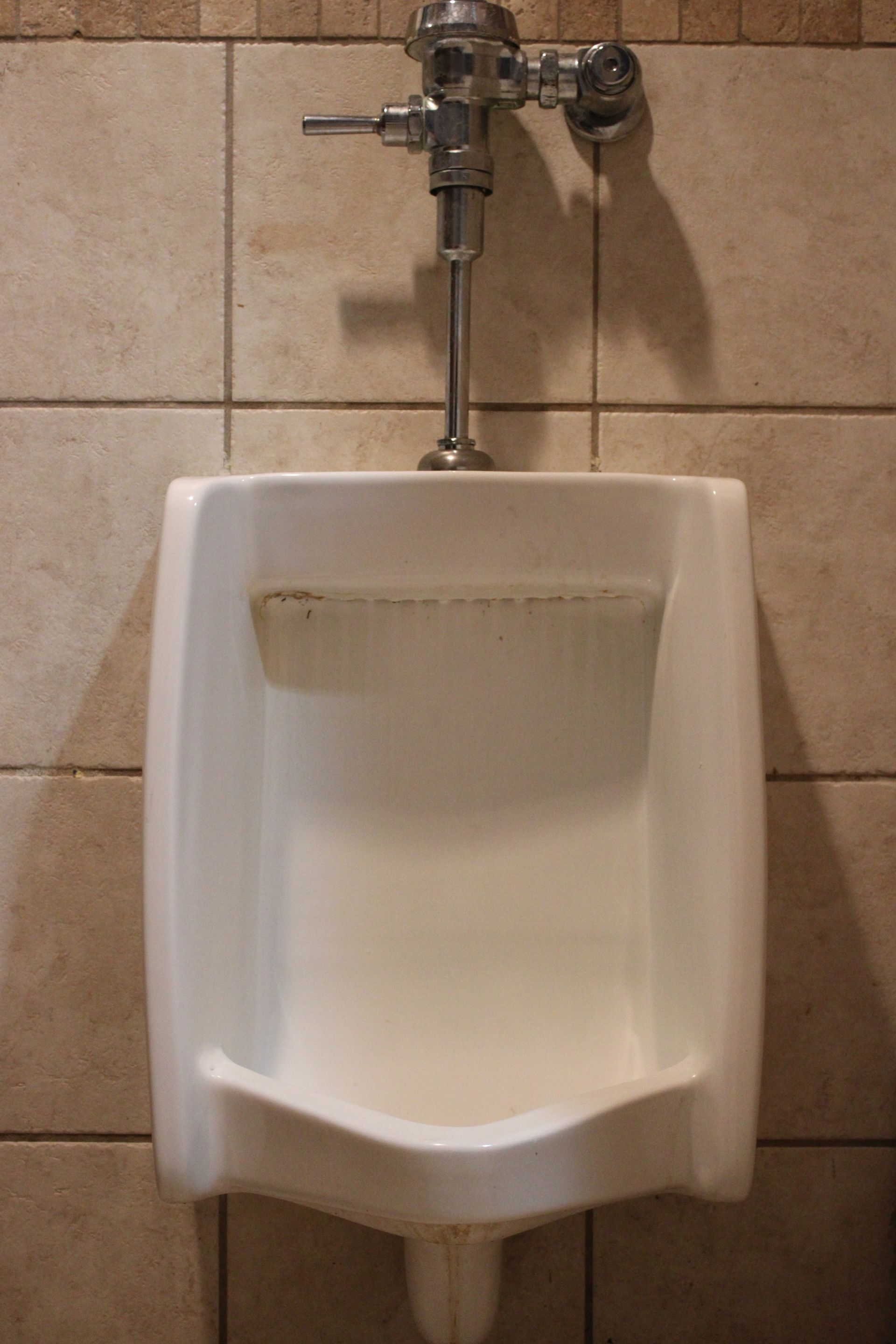 White wall-mounted urinal in a tiled restroom with exposed flush pipes