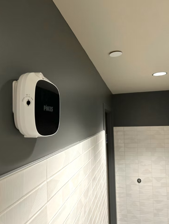 Black and white wall-mounted device in a restroom with gray walls and tiled wainscoting.