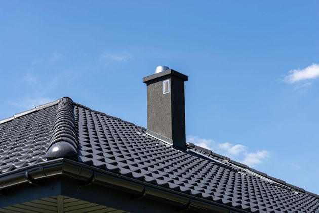 A dark chimney with a metallic cap stands on a black tiled roof against a clear blue sky.