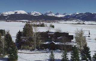An aerial view of a house in the middle of a snowy field with mountains in the background.
