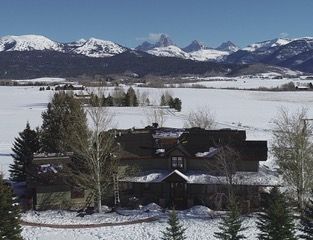There is a house in the middle of a snowy field with mountains in the background.