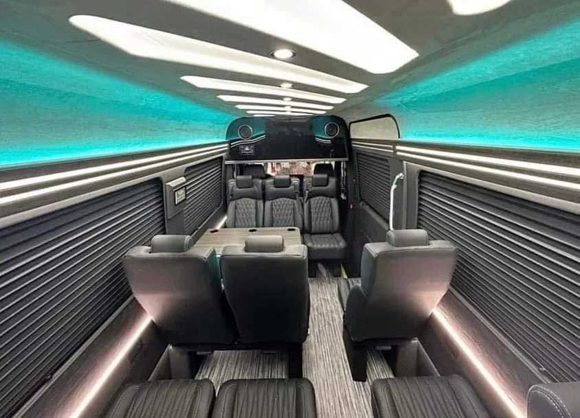 Interior view of a luxury van with black leather seating, wood paneling, and teal lighting along the ceiling.